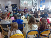 grupa uczniów siedzi przy stolikach w czytelni biblioteki i słucha prelekcji nauczyciela, w tle zdjęcie Sławosza Uznańskiego-Wiśniewskiego ze stacji kosmicznej