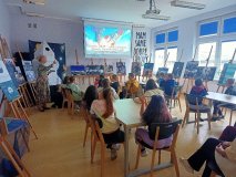 grupa uczniów siedzi przy stolikach w czytelni biblioteki i słucha prelekcji nauczyciela