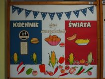 Zdjęcie przedstawia kolorową dekorację przygotowaną z okazji wydarzenia „Kuchnie świata – dzień amerykański”. Na tablicy widnieje napis o tej treści, a całość utrzymana jest w barwach flagi Stanów Zjednoczonych – niebieskiej, białej i czerwonej. U góry zawieszona jest girlanda z niebieskich chorągiewek ozdobionych białymi gwiazdkami. Tablicę wypełniają rysunki typowych potraw kuchni amerykańskiej, takich jak: hamburger, hot dog, frytki i nuggetsy z kurczaka, a także ilustracje warzyw: kukurydzy, papryki i pomidorów.