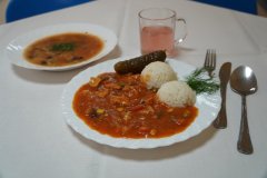 Na zdjęciu widoczny jest zestaw obiadowy na białym obrusie. Na talerzu znajduje się ryż, kurczak w sosie pomidorowym z warzywami oraz ogórek kiszony z gałązką koperku. W tle widać talerz z zupą meksykańską oraz szklankę jasnego kompotu. Obok ułożone są sztućce. 