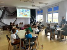 Wnętrze czytelni szkolnej biblioteki, uczniowie 2f siedzą przy stołach i na wiszącym ekranie oglądają prezentację o klockach LEGO. 
