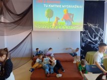 Szkolna biblioteka, na wiszącym ekranie napis „LEGO w bibliotece – tu kwitnie wyobraźnia!”. Na rozłożonym na podłodze materacu leży kilku uczniów i oglądają książkę.