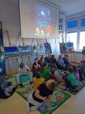 grupa uczniów siedzi lub leży na dywanikach i pufach, słucha audiobooka, w tle ekran, na którym widnieje okładka książki Grażyny Bąkiewicz Kosmiczni odkrywcy