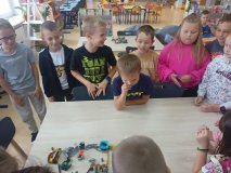 uczniowie przy stołach zgromadzeni w czytelni biblioteki i układający swoje projekty z klocków LEGO