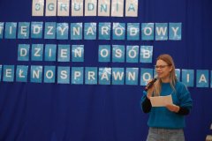 Pani Alicja Wojda i pani Agnieszka Sołtysiak, prowadzące apel z okazji Święta Osób z Niepełnosprawnościami, znajdują się na tle dekoracji z napisem 3 GRUDNIA MIĘDZYNARODOWY DZIEŃ OSÓB Z NIEPEŁNOSPRAWNOŚCIAMI.