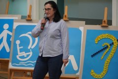 Wicedyrektor Szkoły do spraw integracji i wychowania - pani Iwona Kołodziejska, stoi na tle znaków symbolizujących różne rodzaje niepełnosprawności. Znaki te ustawione są na sztalugach.