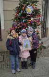 Przed tą samą udekorowaną choinką stoi troje uśmiechniętych Uczniów. Wszyscy mają na sobie ciepłe zimowe ubrania- kurtki, czapki i szaliki. Dzieci pozują razem, stojąc blisko siebie, a ich postawa i mimika pokazują radość. 