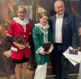 na ciemny brązowym tle ścianki stoją trzy osoby – Prezydent Miasta Płocka Andrzej Nowakowski i dwie uczennice naszej szkoły, obie ubrane w marynarki – jedna ma kolor zielony, druga czerwony, pod szyją mają białe kołnierze stylizowane na renesansowe kryzy