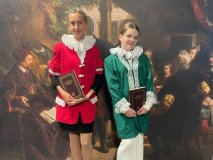 na tle ścianki w kolorze ciemnego brązu stoją dwie uczennice naszej szkoły, obie ubrane w marynarki – jedna ma kolor zielony, druga czerwony, pod szyją uczennic białe kołnierze stylizowane na renesansowe kryzy,dziewczyny w dłoniach trzymają brązowo-złote księgi