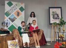 sala Mediateki w Płocku, trzy osoby na scenie z bogatą dekoracją domu szlacheckiego, dwie uczennice siedzą na sofie, obie ubrane w marynarki – jedna ma kolor zielony, druga czerwony, pod szyją mają białe kołnierze stylizowane na renesansowe kryzy, jedna z uczennic ma mikrofon w ręku i czyta utwór, za nimi stoi Wicedyrektor szkoły ubrana w białą bluzkę