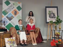 sala Mediateki w Płocku, trzy osoby na scenie z bogatą dekoracją domu szlacheckiego, dwie uczennice siedzą na sofie, patrzą w obiektyw z uśmiechem, za nimi stoi Wicedyrektor szkoły ubrana w białą bluzkę, czyta do mikrofonu