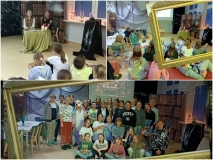 Zbiorowa fotografia uczestników wydarzenia Noc w Bibliotece na tle dekoracji. Z boku przy okrągłym stole siedzą uczennice, z którymi grupa dzieci przeprowadza wywiad na temat pasji i zainteresowań.