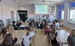 czytelnia biblioteki, uczniowie klasy trzeciej i czwartej siedzą przy stolikach i słuchają prelekcji o nowościach zakupionych w ramach Narodowego Programu Rozwoju Czytelnictwa 2.0. W tle ciemny baner z napisem „Nowości”.