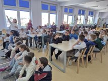 grupa uczniów w czytelni biblioteki siedzi przy stołach, część siedzi na pufach na podłodze, słuchają nauczyciela.