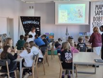 uczniowie w czytelni biblioteki siedzą przy stołach, wśród nich widać trzy nauczycielki pracujące z uczniami, w tle napis „Nowości”.