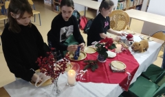 Trzy dziewczynki przygotowują w bibliotece świąteczny stół umieszczając na nim talerze, sztućce, świece, serwetki, gwiazdę betlejemską i inne elementy dekoracyjne.