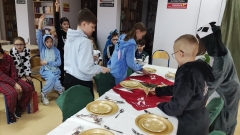 Uczniowie z klasy 4b podczas zajęć w bibliotece przygotowują świąteczny stół umieszczając na nim talerze, sztućce, świece, serwetki, gwiazdę betlejemską i inne elementy dekoracyjne.