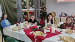 Na tle zimowo-świątecznej dekoracji czworo uczniów z klasy 4b siedzi wokół pięknie udekorowanego stołu.