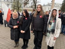 Cztery osoby stoją razem podczas obchodów Narodowego Święta Niepodległości w Płocku. Wszystkie mają przypięte biało-czerwone kotyliony. W tle znajdują się inni uczestnicy wydarzenia oraz widoczne elementy miejskiej zabudowy 