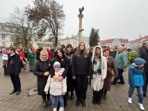 Grupa osób pozuje do wspólnego zdjęcia podczas Obchodów Narodowego Święta Niepodległości na płockim Placu Narutowicza. Wszystkie mają przypięte biało-czerwone kotyliony. W tle widać uczestników uroczystości, flagi oraz kolumnę zwieńczoną orłem 