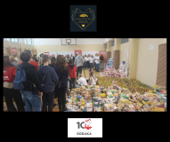 Zdjęcie wykonane w dużej sali gimnastycznej. Na pierwszym planie widoczne są setki paczek z żywnością i napojami ustawionych na podłodze. W tle stoi duża grupa ludzi, głównie młodzież i dorośli, zgromadzonych w kolejce lub grupach. Na ścianach wiszą banery organizacyjne. Całość przedstawia skalę zbiórki i wspólne zaangażowanie wielu osób. Na dole zdjęcia znajduje się logo „10 Paka dla Rodaka”.