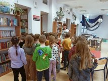 Grupa dzieci w pomieszczeniu biblioteki słucha stoi skupiona wokół bibliotekarki i słucha jej opowieści o zbiorach.