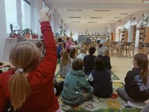 Grupa pierwszoklasistów siedzi na dywanie a bibliotekarz prezentuje im książkę typu pop-up.