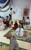 Dzieci siedzą na dywanach a bibliotekarka prezentuje na ścianie książkę „Gra cieni” – podświetla latarką ażurowe strony książki, które rzucają cienie na ścianę.