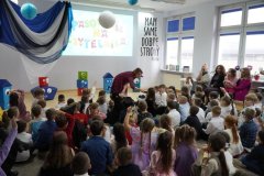 Uroczystość pasowania na czytelnika, grupa pierwszaków siedzi na podłodze a bibliotekarka podsuwa jednemu z dzieci mikrofon, by mogło udzielić odpowiedzi na pytanie.