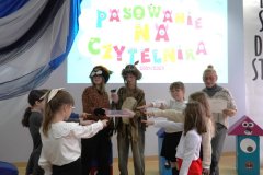 Moment pasowania – dwie siódmoklasistki w brązowym przebraniu jako Pożeracze Książek, trzymają książkę. Sześcioro pierwszoklasistów stoi wokół księgi z bajkami z wyciągniętymi w jej stronę dwoma palcami prawej ręki i składają przyrzeczenie.