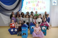 Zdjęcie grupowe klasy 1b, uczniowie pozują w kolorowych bajkowych strojach, wokół nich figury zabawnych potworków.