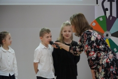 Uczeń klasy pierwszej podaje do mikrofonu odpowiedź na zagadkę zadaną mu w ramach turnieju „Koło fortuny”, w tle inni uczniowie