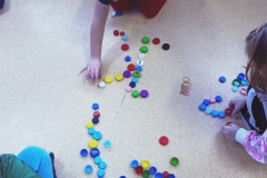 Pierwszaki układają z plastikowych korków od butelek pierwszą literę swojego imienia – widok na trzy osoby