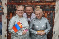 Trzy osoby pozują do zdjęcia na tle wzorzystej tkaniny; jedna z kobiet trzyma małą flagę Mongolii.