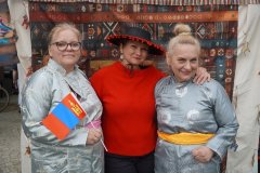 Trzy kobiety pozują przed namiotem; dwie mają na sobie tradycyjne stroje, a jedna trzyma flagę Mongolii.