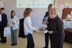Dyrektor Szkoły Podstawowej nr 22 i przewodnicząca jury wręczają dziewczynce dyplom i torbę z logo konkursu, w tle widać innych laureatów