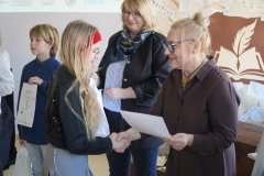 Dyrektor Szkoły Podstawowej nr 22 i przewodnicząca jury wręczają dziewczynce dyplom i torbę z logo konkursu, w tle widać innych laureatów