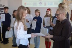 Dyrektor Szkoły Podstawowej nr 22 i przewodnicząca jury wręczają dziewczynce dyplom i torbę z logo konkursu, w tle widać innych laureatów