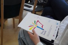 zbliżenie na publikację, która zawiera fragmenty nagrodzonych archiwalnych prac, na okładce logo konkursu – kolorowe pióro, leżącą na kolanach siedzącej uczestniczki konkursu