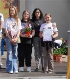 foto_01c grupa czterech uśmiechniętych osób, w tym trzy dziewczynaki i dorosła kobieta, pozują wspólnie na scenie podczas uroczystości na świeżym powietrzu. Jedno z dzieci trzyma dyplom, drugie roślinę doniczkową