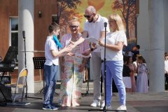 foto_01d chłopiec odbiera gratulacje i dokument od dorosłych podczas uroczystości na scenie plenerowej. Obok niego stoją trzy osoby dorosłe, a w tle widoczna jest grupa dzieci i dekoracja z motywem rodzinnym
