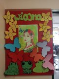 foto_13 plakat: na czerwonym tle ilustracja pięknej wiosny w postaci kobiety, wokół której krążą motyle i znajdują sie kolorowe kwiaty i zielone żabki wyciete z papieru