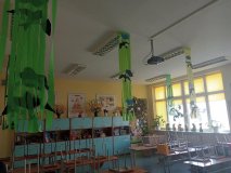 foto_8 sala lekcyjna, w której przy suficie zostały zawieszone kurtynki z zielonych pasków z bibuły, do których zostały przyklejone wycięte z kolorowego paieru jaskółki i chmurki, w w tle okien duże ażurowe motyle, a na parapecie włąsnoręcznie wykonane wiosenne stroiki