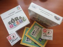 Zestaw nowych gier i publikacji terapeutycznych na drewnianym blacie. Na zdjęciu widoczne są pudełka z grami „Góra Uczuć” oraz „Gra na emocjach”, a także książki z serii „Gry terapeutyczne dla nastolatków”.