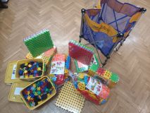 estaw nowych zabawek i pomocy dydaktycznych na drewnianym parkiecie. Na zdjęciu widać kolorowe klocki typu wafle w opakowaniach i pudełkach oraz rozkładany, niebiesko-żółty kosz z siatki na akcesoria.