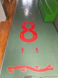 Czerwona naklejka podłogowa na korytarzu przedstawiająca dużą cyfrę osiem ze strzałkami pomocniczymi do nauki kierunku kreślenia lub poruszania się. Poniżej widoczna jest falista linia z rakami oraz strzałki i ślady stóp prowadzące w głąb korytarza.