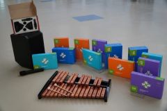 Zestaw nowoczesnych pomocy dydaktycznych na jasnej podłodze. Na pierwszym planie drewniane dzwonki (ksylofon) z pałeczkami, obok głośnik z mikrofonem, a w tle rząd kolorowych pudełek z puzzlami edukacyjnymi przeznaczonymi do pracy z robotami Ozobot.