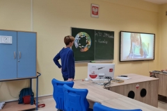 Nauczyciel stoi przy biurku, na tradycyjnej tablicy obrazek puzzli, na interaktywnej - postać dziewczynki.