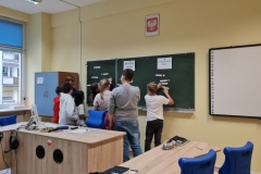 Nauczyciel z grupą dzieci stoją przy tablicy i wspólnie układają zadanie.
