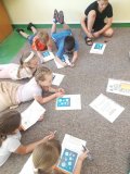 Uczniowie siedzący swobodnie na dywanie wykonują zadania na kartach  pracy.